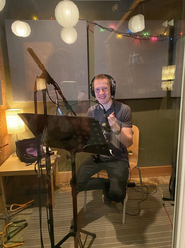 Aleh Remezau in a recording booth wearing headphones, oboe in hand, in front of a music stand and microphone